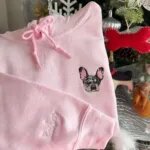 Light pink hoodie with an embroidered merle French bulldog face on the chest and a script name on the sleeve