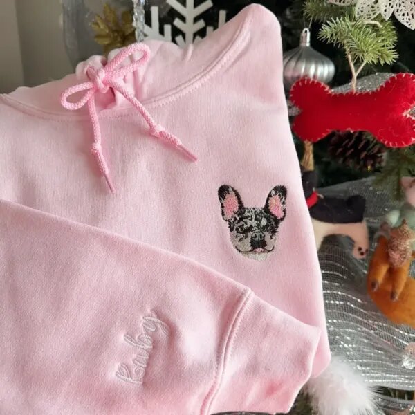 Light pink hoodie with an embroidered merle French bulldog face on the chest and a script name on the sleeve