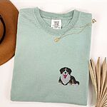 Bay T-shirt laid flat with a necklace and dried palm leaves, embroidered with a small tricolor Berner puppy lying down on the left chest