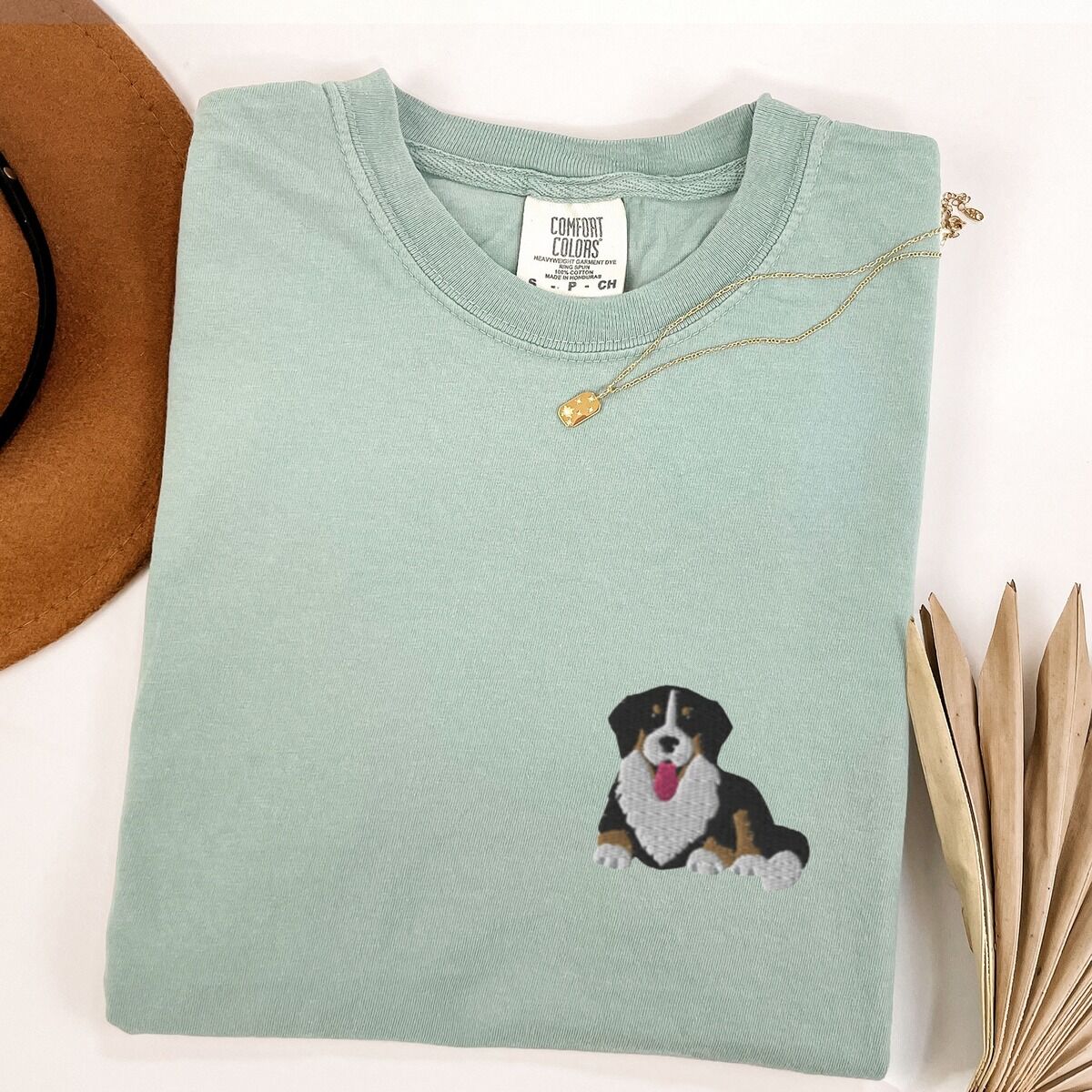 Cute Bernese Mountain Dog Shirt – Bay Bay T-shirt laid flat with a necklace and dried palm leaves, embroidered with a small tricolor Berner puppy lying down on the left chest