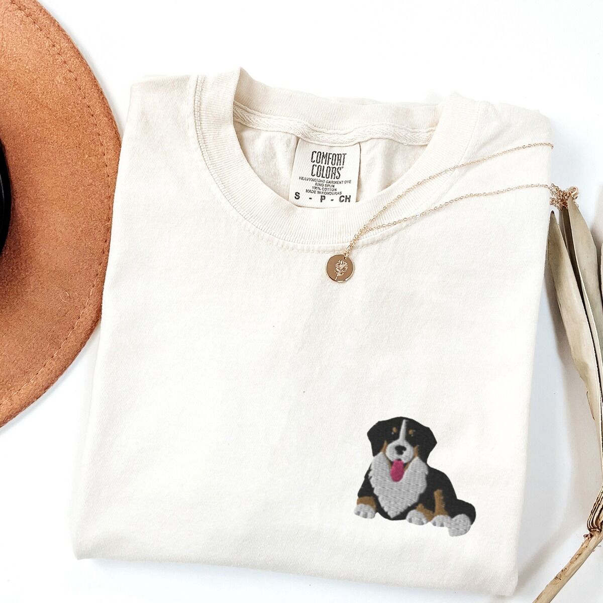 Cute Bernese Mountain Dog Shirt – Ivory Ivory T-shirt laid flat with a necklace and a felt hat, embroidered with a cute Bernese mountain dog puppy lying down on the left chest