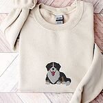 Sand crewneck sweatshirt laid flat on herringbone wood, embroidered with a larger Bernese mountain dog puppy lying down in the center of the chest