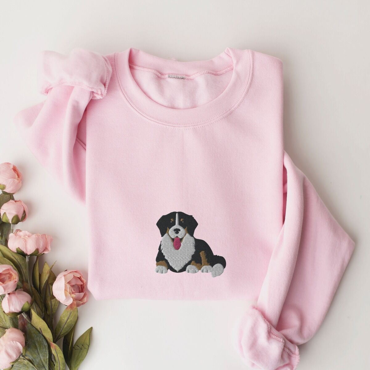 Cute Bernese Mountain Dog Sweatshirt - center chest in pink Light Pink crewneck sweatshirt laid flat with pink roses, embroidered with a larger Bernese mountain dog puppy with its tongue out in the center of the chest