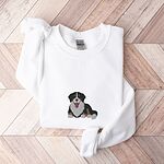 White crewneck sweatshirt laid flat on herringbone wood, embroidered with a larger Berner puppy lying down in the center of the chest
