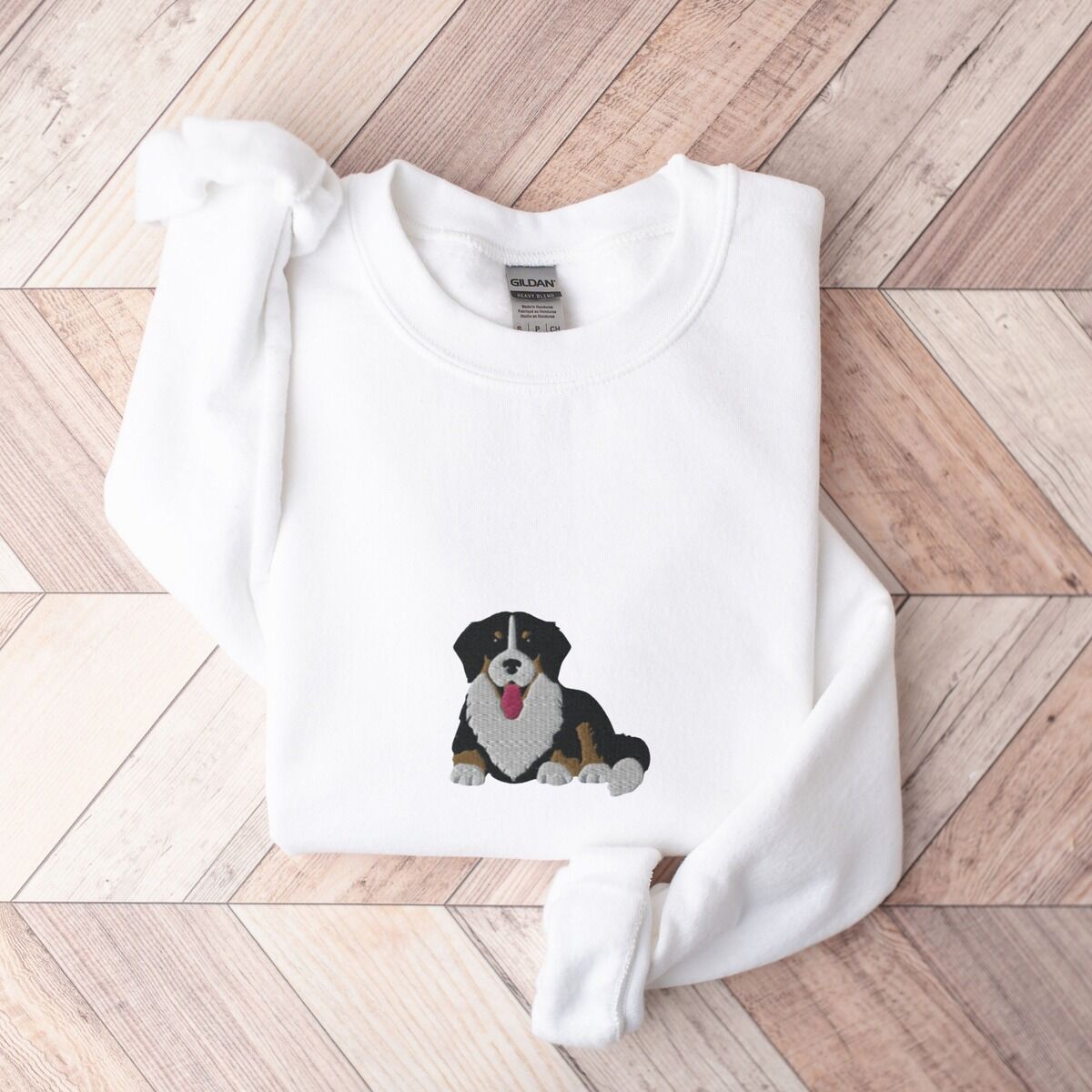 Cute Bernese Mountain Dog Sweatshirt - center chest in white White crewneck sweatshirt laid flat on herringbone wood, embroidered with a larger Berner puppy lying down in the center of the chest