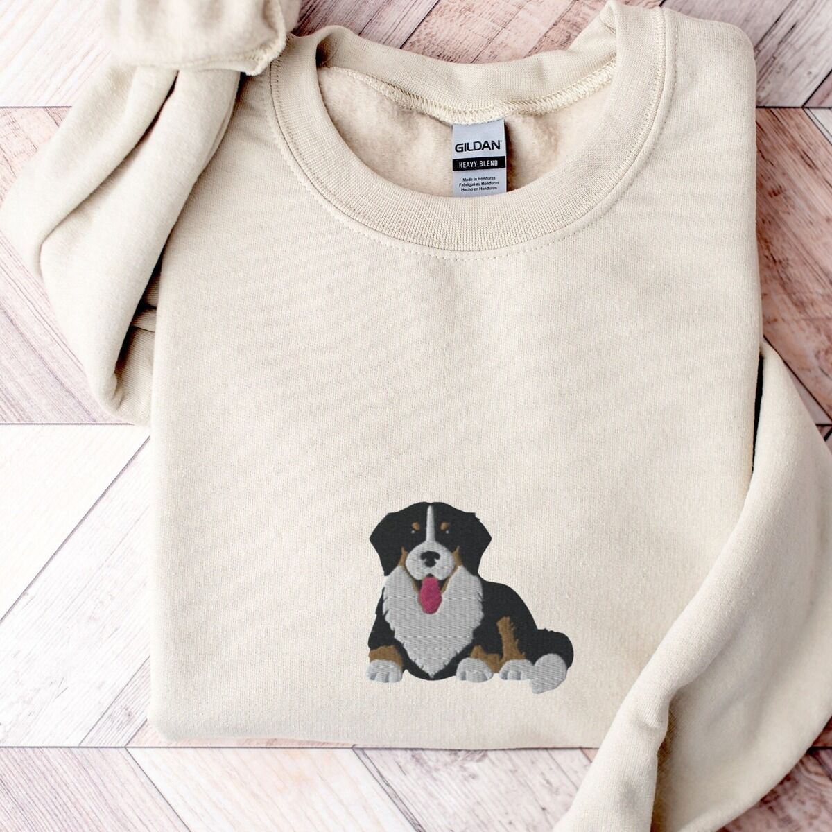 Cute Bernese Mountain Dog Sweatshirt - center chest Sand crewneck sweatshirt laid flat on herringbone wood, embroidered with a larger Bernese mountain dog puppy lying down in the center of the chest