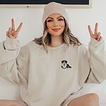 Woman in a beanie making peace signs in a Sand crewneck sweatshirt embroidered with a small Berner puppy on the left chest