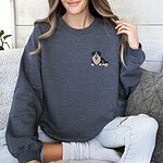 Woman on a sofa in a Dark Heather crewneck sweatshirt embroidered with a small Bernese mountain dog puppy lying down on the left chest