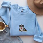 Light Blue crewneck sweatshirt laid flat with a coffee cup, jeans and a felt hat, embroidered with a larger Berner puppy in the center of the chest