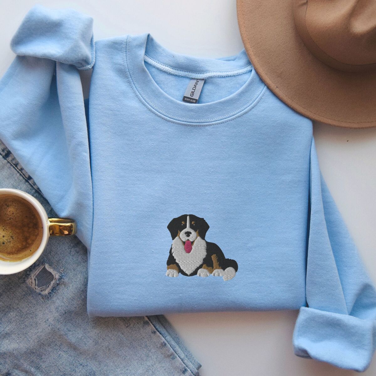 Cute Bernese Mountain Dog Sweatshirt - Light Blue Light Blue crewneck sweatshirt laid flat with a coffee cup, jeans and a felt hat, embroidered with a larger Berner puppy in the center of the chest