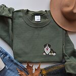 Military Green crewneck sweatshirt laid flat with a felt hat, jeans and autumn leaves, embroidered with a small tricolor Berner puppy on the left chest