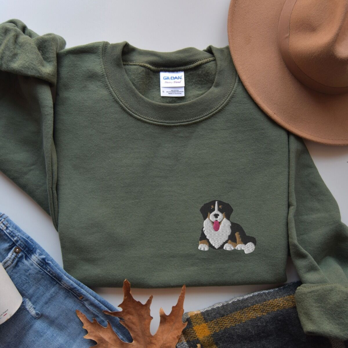 Cute Bernese Mountain Dog Sweatshirt – Military Green Military Green crewneck sweatshirt laid flat with a felt hat, jeans and autumn leaves, embroidered with a small tricolor Berner puppy on the left chest