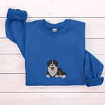 Royal crewneck sweatshirt laid flat on whitewashed wood, embroidered with a larger tricolor Bernese mountain dog puppy in the center of the chest
