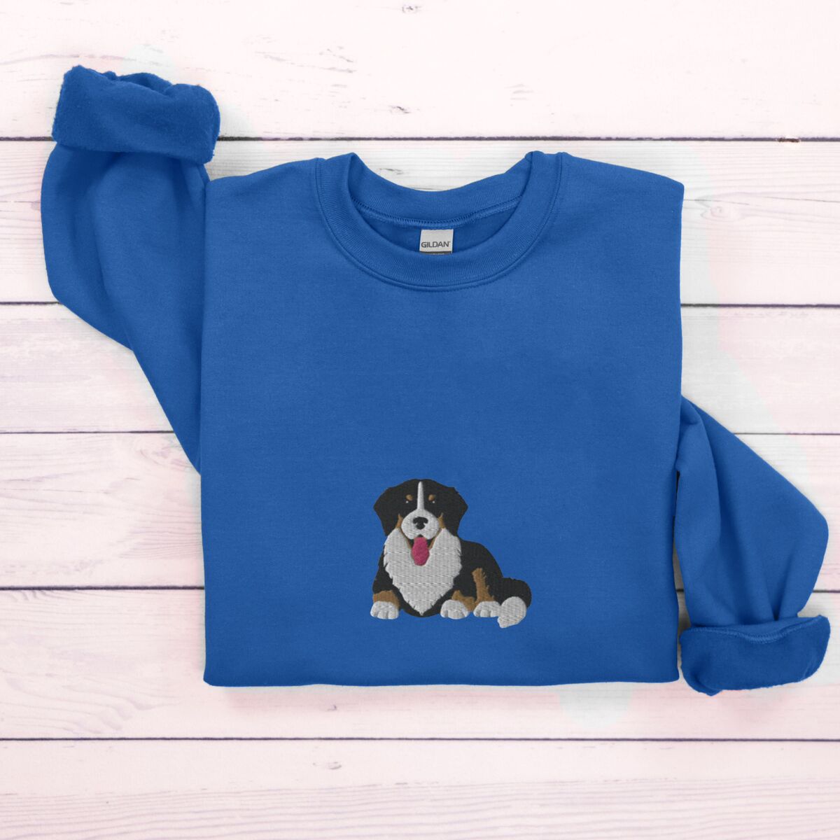 Cute Bernese Mountain Dog Sweatshirt - Royal Royal crewneck sweatshirt laid flat on whitewashed wood, embroidered with a larger tricolor Bernese mountain dog puppy in the center of the chest