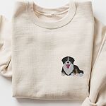 Sand crewneck sweatshirt laid flat on white, embroidered with a small cute Bernese mountain dog puppy lying down with its tongue out on the left chest