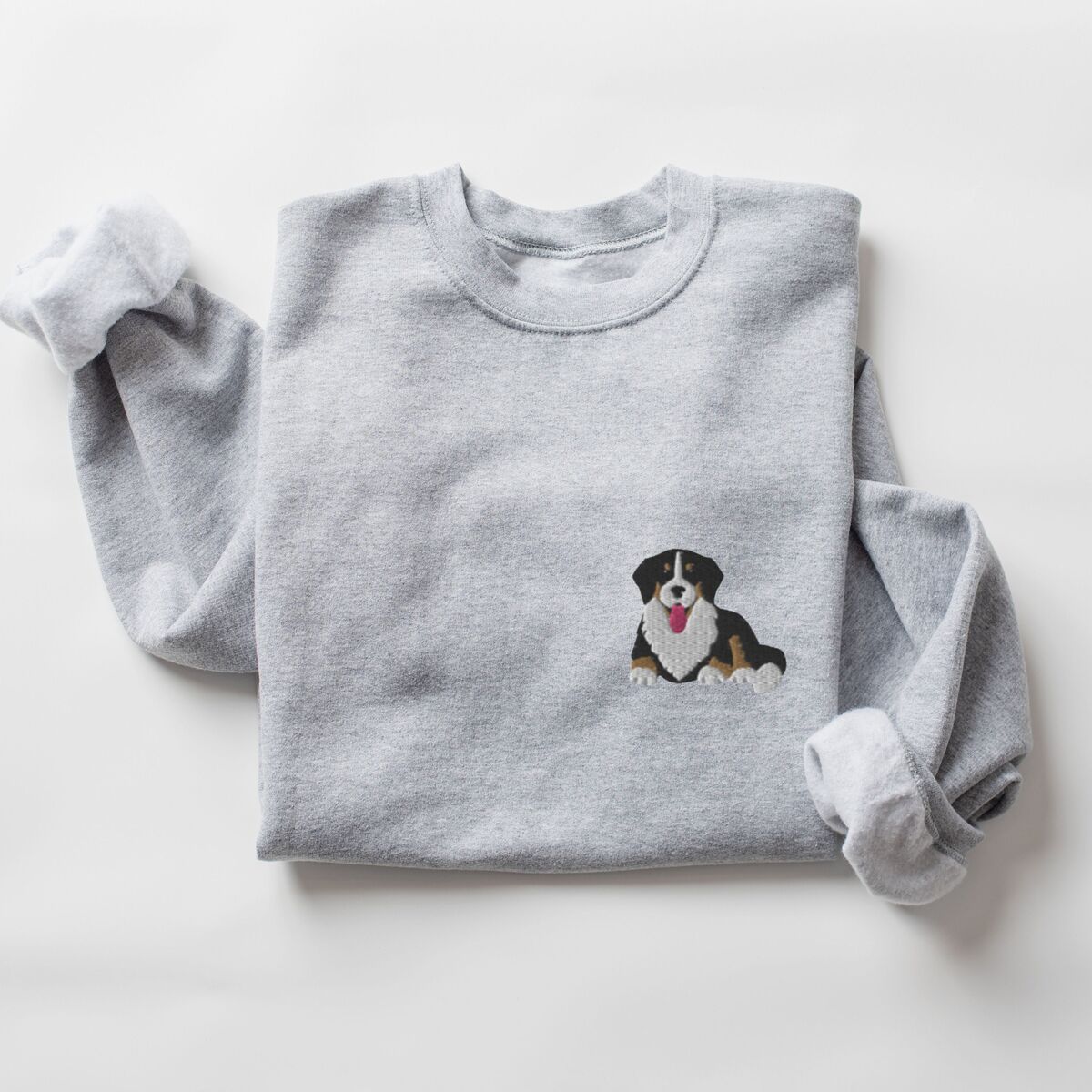 Cute Bernese Mountain Dog Sweatshirt - Sport Grey Sport Grey crewneck sweatshirt laid flat on white, embroidered with a small Berner puppy lying down on the left chest