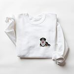 White crewneck sweatshirt laid flat on white, embroidered with a small tricolor Bernese mountain dog puppy lying down with its tongue out on the left chest