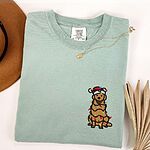 Bay T-shirt laid flat with a necklace, embroidered with a cartoon golden retriever in a Santa hat wrapped in Christmas lights on the left chest