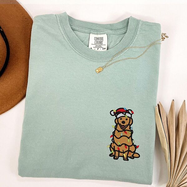 Bay T-shirt laid flat with a necklace, embroidered with a cartoon golden retriever in a Santa hat wrapped in Christmas lights on the left chest