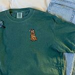 Blue Spruce T-shirt laid flat beside jeans and glasses, embroidered with a cartoon golden retriever wrapped in Christmas lights on the left chest