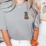Woman in a light grey T-shirt embroidered with a cartoon golden retriever in a Santa hat tangled in Christmas lights on the left chest