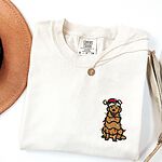 Ivory T-shirt laid flat with a necklace, embroidered with a cartoon golden retriever in a Santa hat and a string of colored lights