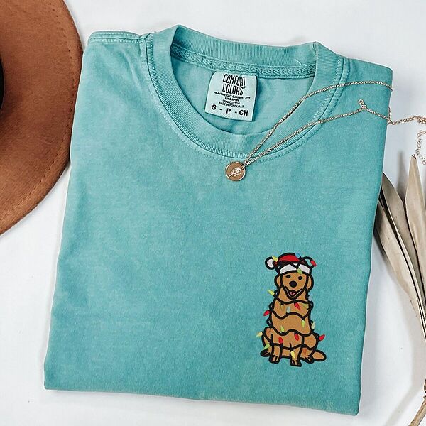 Cute golden retriever Christmas shirt in Seafoam laid flat with a necklace and a straw hat, embroidered with a cartoon golden in a Santa hat tangled in Christmas lights
