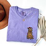 Violet T-shirt laid flat with a necklace, embroidered with a smiling cartoon golden retriever tangled in Christmas lights