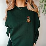 Woman in a Forest Green crewneck sweatshirt embroidered with a cartoon golden retriever in a Santa hat wrapped in Christmas lights