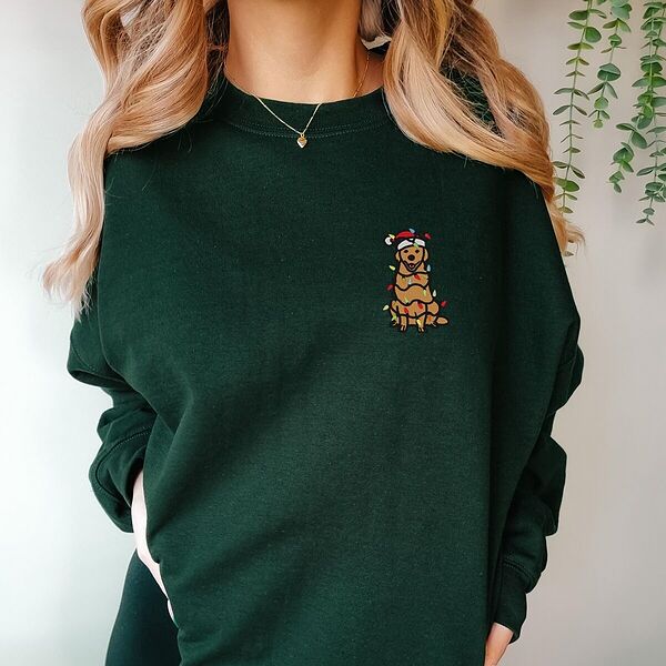 Woman in a Forest Green crewneck sweatshirt embroidered with a cartoon golden retriever in a Santa hat wrapped in Christmas lights