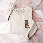 Sand crewneck sweatshirt laid flat on a wood floor, embroidered with a smiling cartoon golden retriever wrapped in Christmas lights