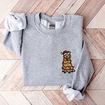 Sport Grey crewneck sweatshirt laid flat on a wood floor, embroidered with a cartoon golden retriever in a Santa hat tangled in lights