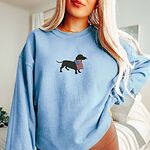 Woman in a Light Blue crewneck sweatshirt with a black dachshund holding a USA flag centered on the chest