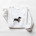 White crewneck sweatshirt laid flat, embroidered with a black doxie carrying a red, white and blue flag