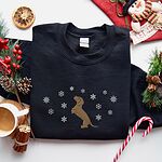 Black crewneck sweatshirt laid flat beside a Christmas stocking, a mug of coffee and a candy cane, embroidered with a dachshund standing up on its hind legs with its nose to the sky among falling snowflakes