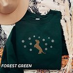 Forest Green crewneck sweatshirt laid flat with a felt hat and pampas grass, embroidered with a dachshund standing up on its hind legs with its nose to the sky among falling snowflakes
