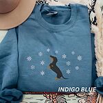 Indigo Blue crewneck sweatshirt laid flat with a felt hat and pampas grass, embroidered with a dachshund standing up on its hind legs with its nose to the sky among falling snowflakes