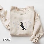 Sand crewneck sweatshirt folded on a white background, embroidered with a dachshund standing up on its hind legs with its nose to the sky among falling snowflakes