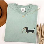 Bay T-shirt laid flat with a necklace and a straw hat, embroidered with a black and tan dachshund trotting with a string of colored Christmas lights in its mouth on the left chest