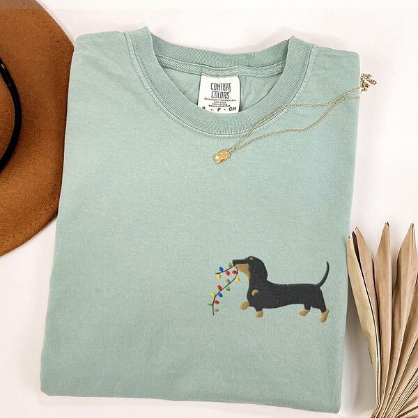 Bay T-shirt laid flat with a necklace and a straw hat, embroidered with a black and tan dachshund trotting with a string of colored Christmas lights in its mouth on the left chest