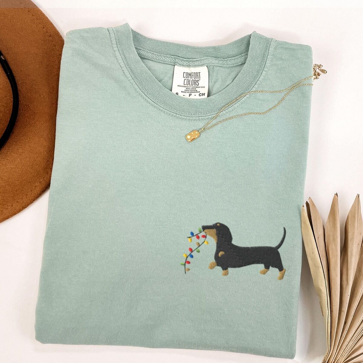 Dachshund Christmas Lights Shirt – Bay Bay T-shirt laid flat with a necklace and a straw hat, embroidered with a black and tan dachshund trotting with a string of colored Christmas lights in its mouth on the left chest