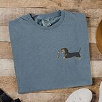Blue Jean T-shirt laid flat on wood beside white sneakers, embroidered with a black and tan dachshund trotting with a string of colored Christmas lights in its mouth on the left chest