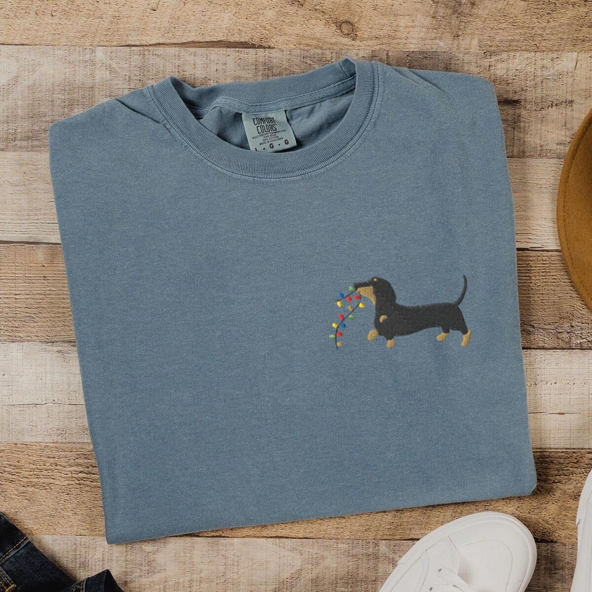 Dachshund Christmas Lights Shirt – Blue Jean Blue Jean T-shirt laid flat on wood beside white sneakers, embroidered with a black and tan dachshund trotting with a string of colored Christmas lights in its mouth on the left chest