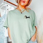Woman in a pale green T-shirt, embroidered with a black and tan dachshund trotting with a string of colored Christmas lights in its mouth on the left chest