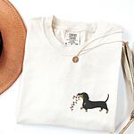 Ivory T-shirt laid flat with a necklace and a straw hat, embroidered with a black and tan dachshund trotting with a string of colored Christmas lights in its mouth on the left chest