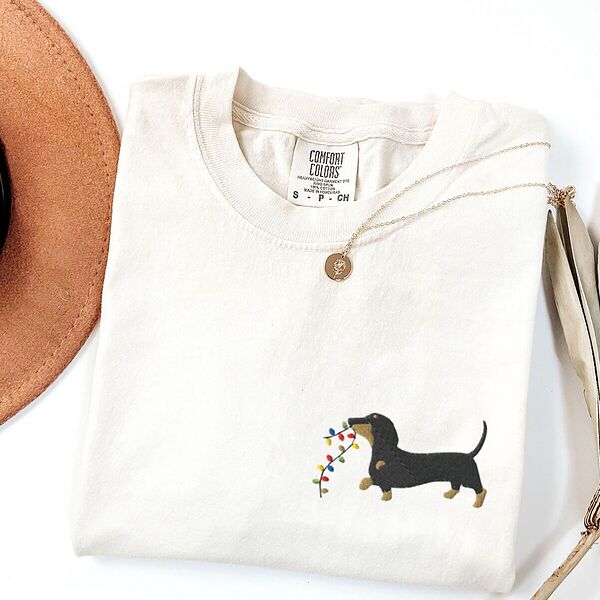Ivory T-shirt laid flat with a necklace and a straw hat, embroidered with a black and tan dachshund trotting with a string of colored Christmas lights in its mouth on the left chest