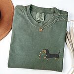 Moss T-shirt laid flat with a necklace and a straw hat, embroidered with a black and tan dachshund trotting with a string of colored Christmas lights in its mouth on the left chest