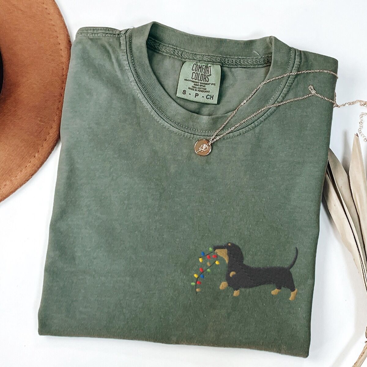 Dachshund Christmas Lights Shirt – Moss Moss T-shirt laid flat with a necklace and a straw hat, embroidered with a black and tan dachshund trotting with a string of colored Christmas lights in its mouth on the left chest