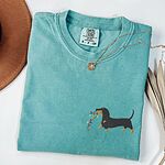 Seafoam T-shirt laid flat with a necklace and a straw hat, embroidered with a black and tan dachshund trotting with a string of colored Christmas lights in its mouth on the left chest