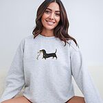 Smiling woman sitting in a light grey crewneck sweatshirt, embroidered with a black and tan dachshund trotting with a string of colored Christmas lights in its mouth